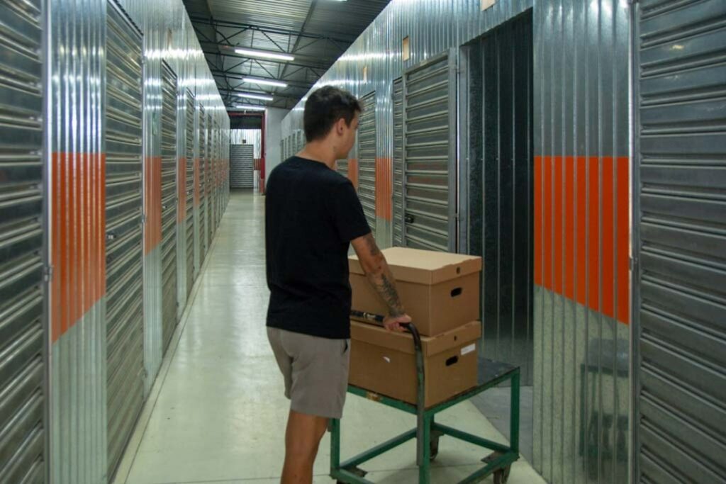 Homem empurra carrinho com caixas de papel&atilde;o em corredor de self storage com boxes met&aacute;licos.